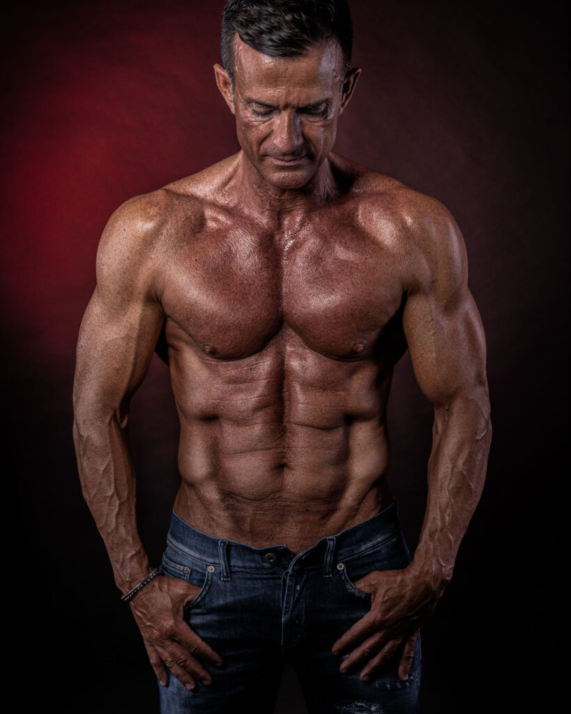 fotografo fitness servizio fotografico in studio con sfondo rosso e ragazzo muscoloso a torso nudo con addominale scolpito e blue jeans